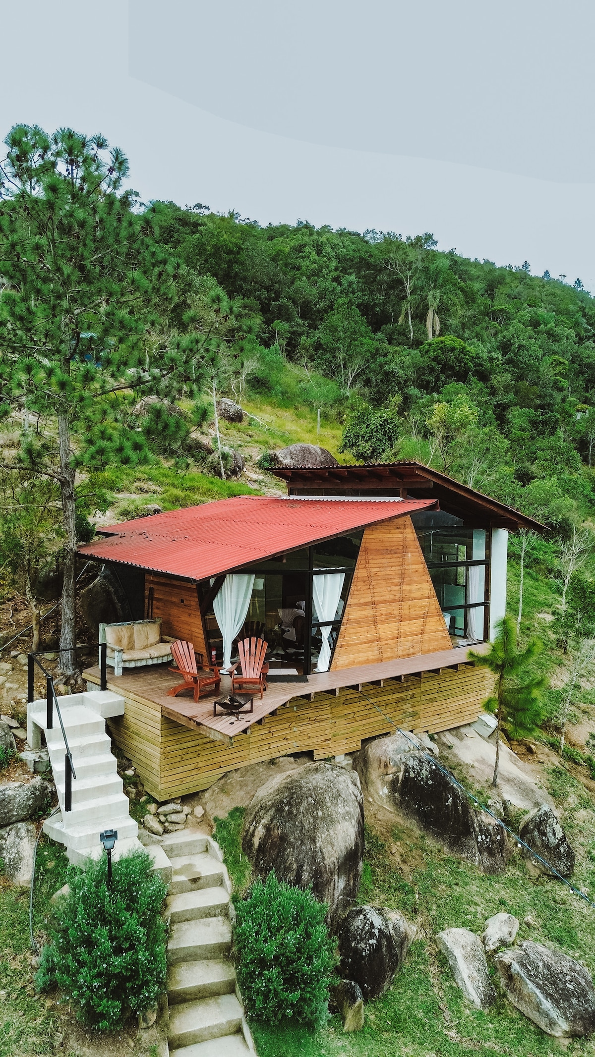 A cabin features a unique architectural design with wooden siding and large glass windows. The outdoor area includes seating and is surrounded by greenery and rocky terrain, providing a natural setting. A staircase leads to the entrance, blending the cabin with the landscape.