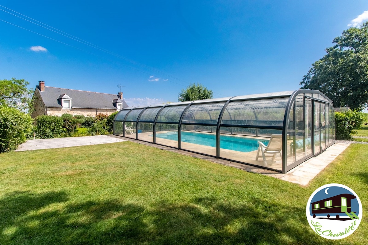 An enclosed swimming pool is framed by a well-maintained lawn and hedges. The pool area is protected by a clear, curved glass structure, allowing sunlight to illuminate the water. In the background, a rustic building is visible against a clear blue sky.