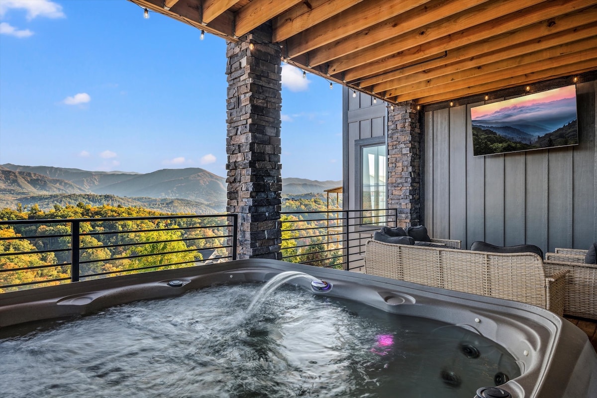 A hot tub equipped for six is placed on a covered terrace, surrounded by stone walls. Views of lush mountains are visible in the background. An outdoor television is mounted on the wall, providing entertainment. Soft lighting enhances the space's ambiance.