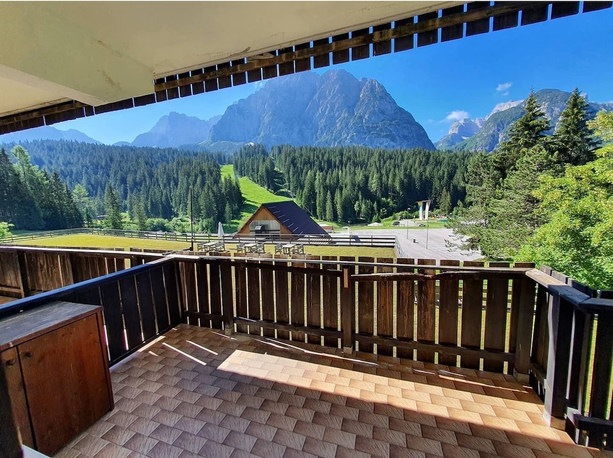 A spacious balcony offers stunning mountain views, surrounded by lush greenery. The floor is tiled with light-colored ceramic tiles. Wooden railings frame the area, providing a cozy yet open feel. The peaceful landscape includes a nearby chalet and expansive forested hills.
