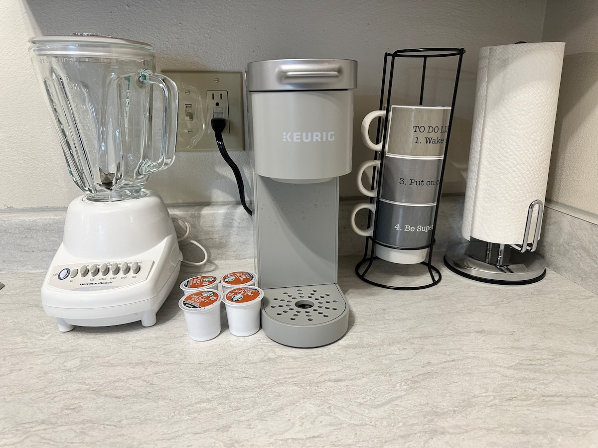A countertop features a Keurig coffee maker alongside a blender and a set of coffee mugs. A roller holder is present, containing paper towels. Coffee pods are arranged neatly in front of the coffee maker, providing convenient options for brewing.