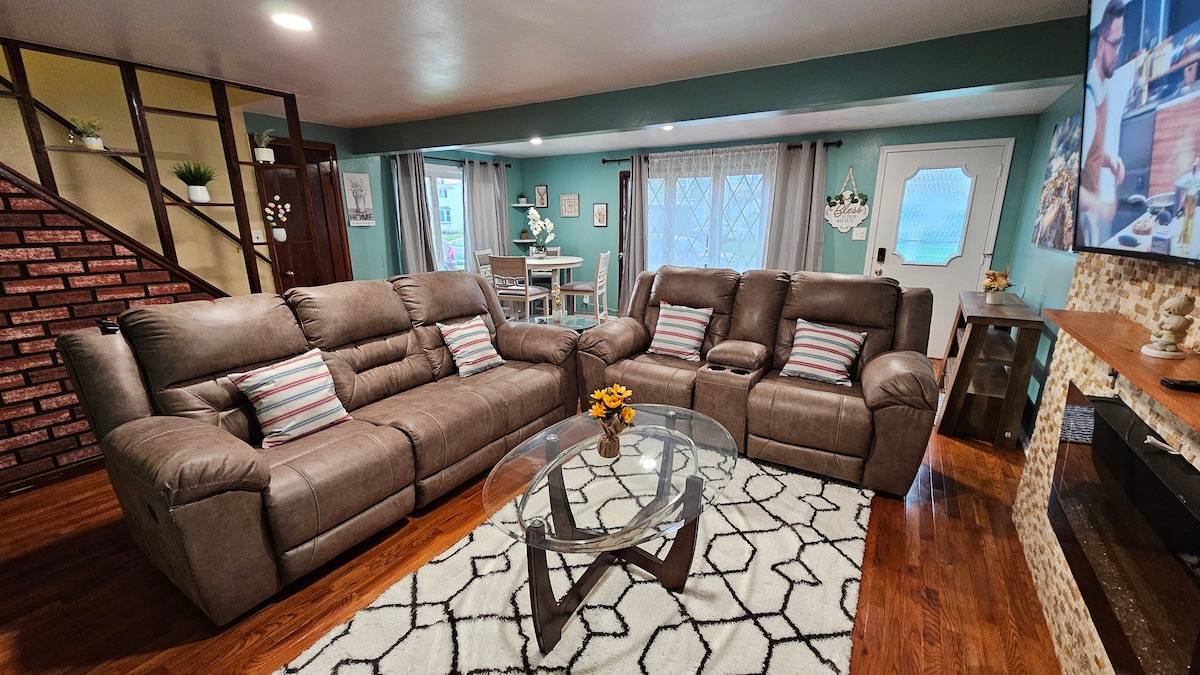 A cozy living room area features a large, comfortable sectional sofa with throw pillows, positioned around a circular glass coffee table. To the left, a staircase leads to upper levels, while a dining area is visible in the background, illuminated by natural light from large windows.