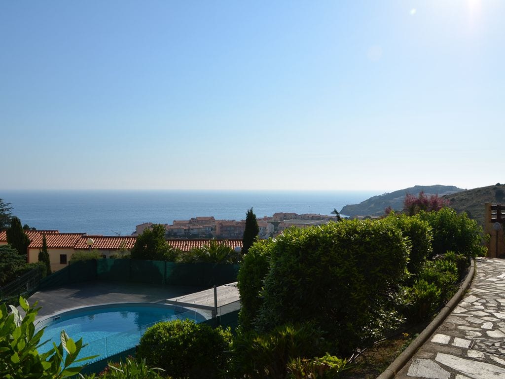 A panoramic view of the Mediterranean coastline is presented, featuring a shared swimming pool edged with greenery. The gentle slope of the landscape leads down to coastal buildings, while the clear sky complements the serene atmosphere.