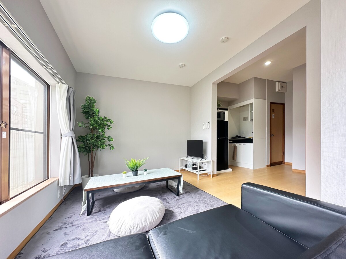 A bright living area features large windows allowing natural light to flow in. A green potted plant adds a touch of nature. A low coffee table sits on a soft rug next to a comfortable sectional sofa, with a television visible in the corner.