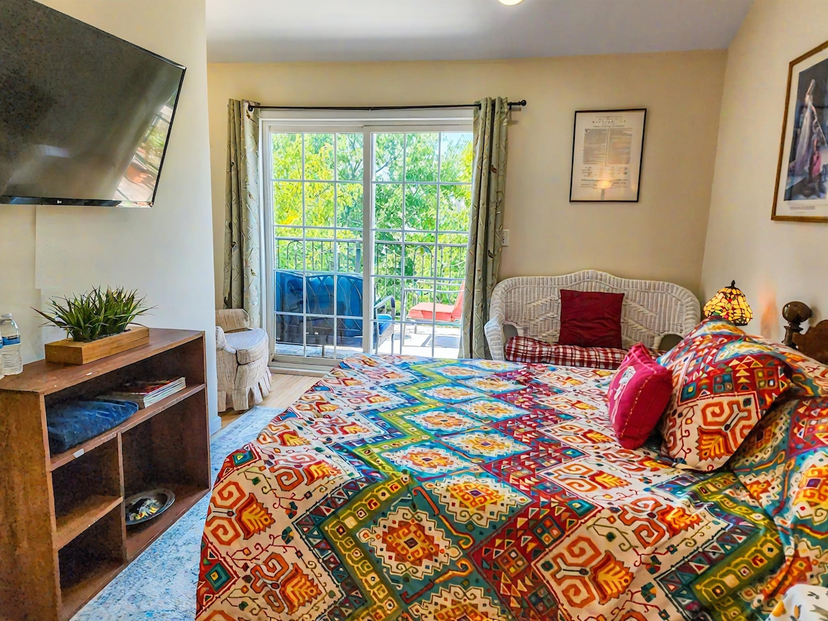 A cozy bedroom features a colorful quilted bedspread with intricate patterns. Natural light fills the room through glass doors leading to a private balcony. A mounted television is visible on the wall, and a small seating area with a light-colored couch adds comfort to the space.