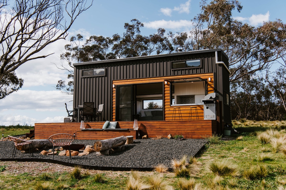A modern Tiny House is presented amidst natural surroundings, featuring a wooden deck with seating and a fire pit area. Large windows invite light into the interior, while rustic logs are arranged decoratively on the deck, enhancing the connection to nature.