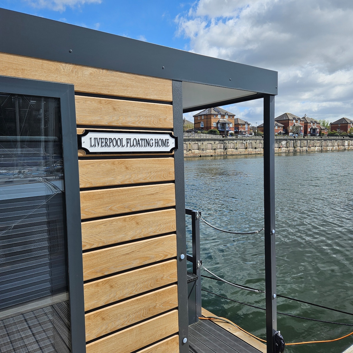 The exterior of the floating home is shown, featuring a wooden facade with a neatly affixed sign indicating 'Liverpool Floating Home.' The surrounding waterfront offers a view of the marina and nearby buildings under a partly cloudy sky.