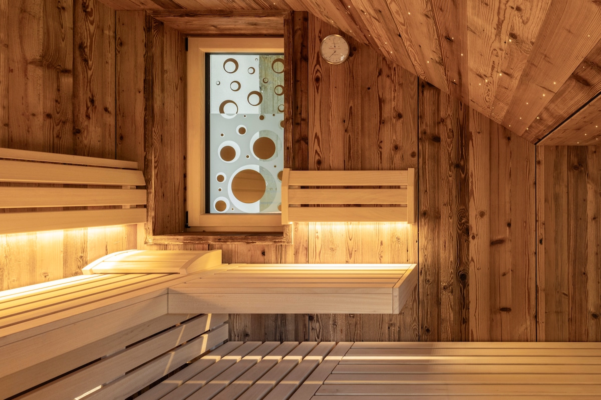 A wooden sauna features benches arranged along the walls, illuminated by soft lighting. A circular window allows natural light to enter, revealing a unique design with various sizes of circles. The warm wood tones contribute to a serene and inviting atmosphere.