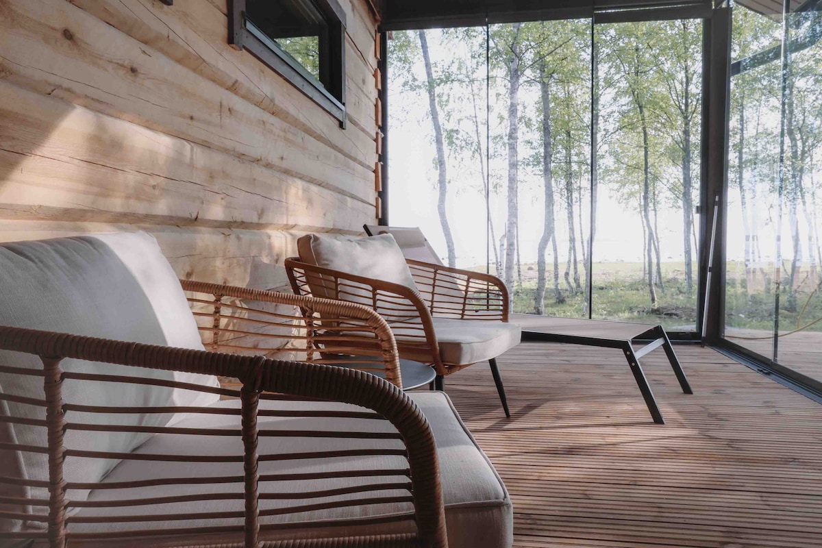 A cozy seating area features two cushioned armchairs made of natural materials, positioned beside a sleek, minimalistic table. Floor-to-ceiling glass walls provide unobstructed views of lush greenery outside, immersing the space in natural light.