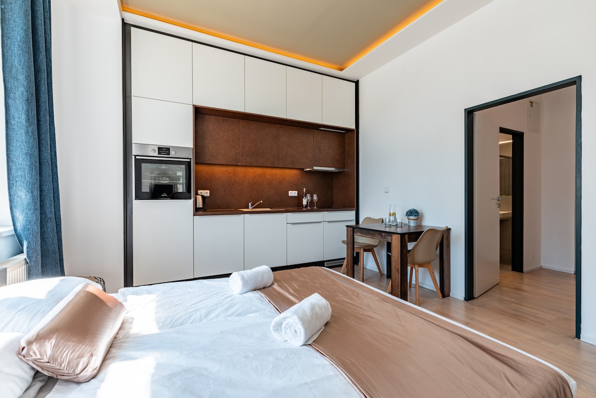 A modern studio layout is displayed, featuring a spacious bed with neatly arranged linens and towels. A kitchen area is equipped with contemporary amenities. A small dining table with two chairs is placed beside the bed, and warm lighting accents the ceiling.