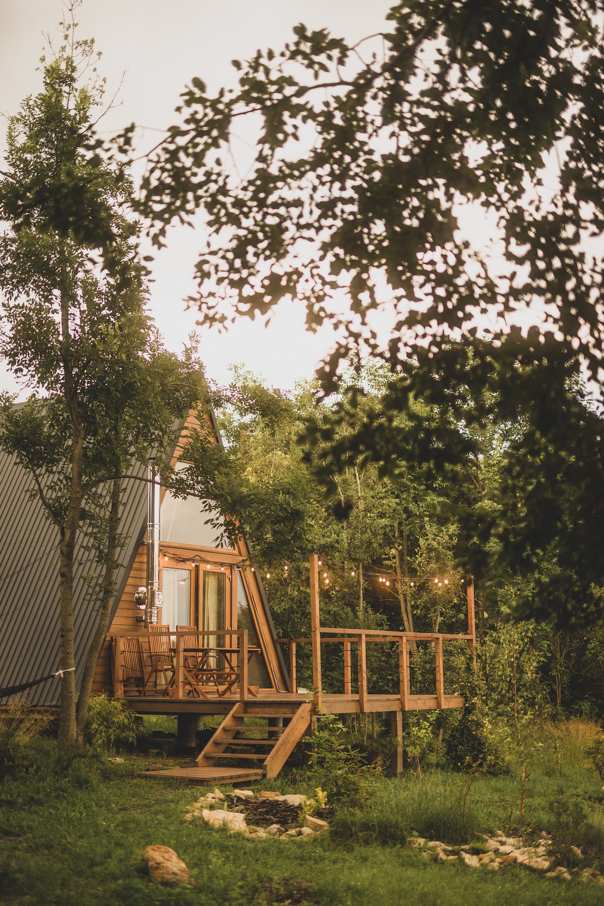 A wooden cabin is nestled among lush greenery, featuring a deck area with outdoor seating. Soft lighting strands are seen above the deck, adding to the serene outdoor atmosphere. A stone fire pit is visible in the foreground, surrounded by natural landscaping.
