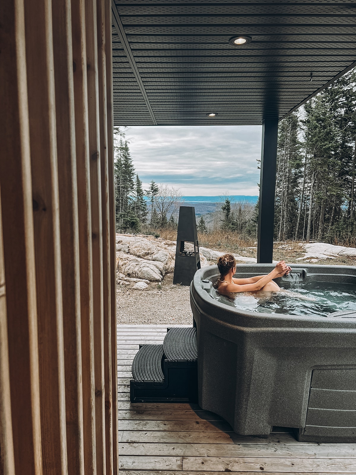 A hot tub is situated on a wooden deck, surrounded by nature. A person is relaxing in the water, while trees and distant hills are visible in the background. The atmosphere is serene, inviting guests to unwind and enjoy the tranquil views.