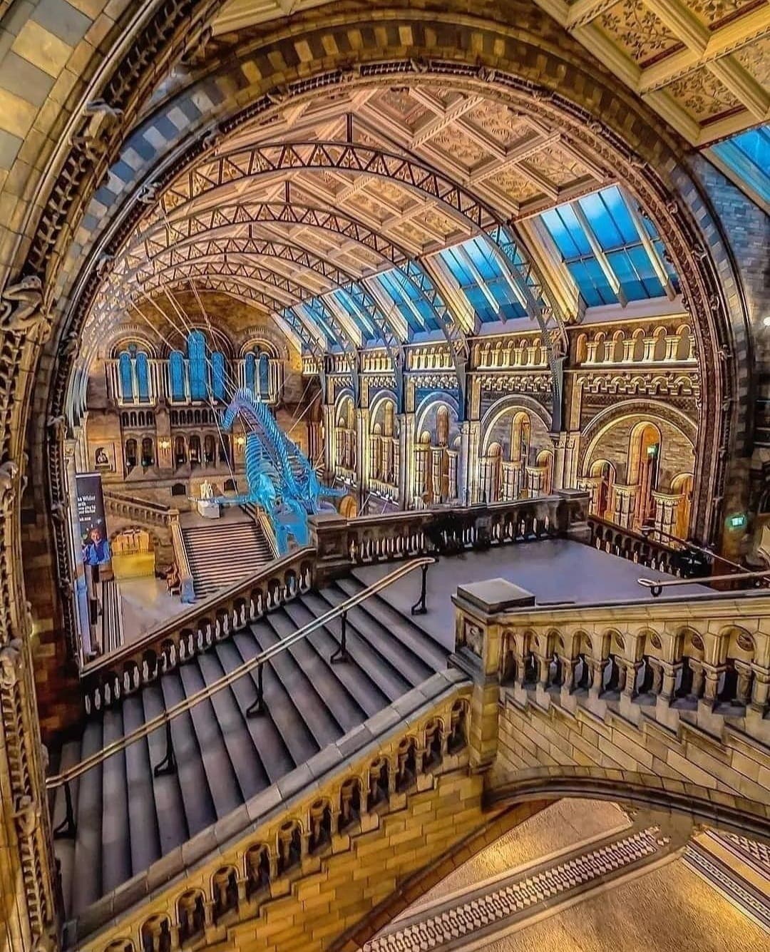 The grand staircase leads to an expansive hall with intricate architectural details. A stunning ceiling features large glass panels allowing natural light to illuminate the space. Ornate columns and arches enhance the historical ambiance, while a striking blue installation draws the eye.