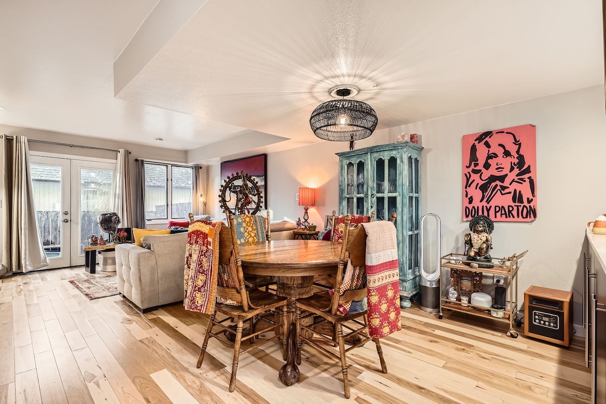 A cozy dining area features a round wooden table surrounded by rustic chairs, complemented by a colorful table runner. A vibrant artwork of Dolly Parton adds character to the wall. Natural light filters through large windows, enhancing the inviting ambiance of the open-concept living space.