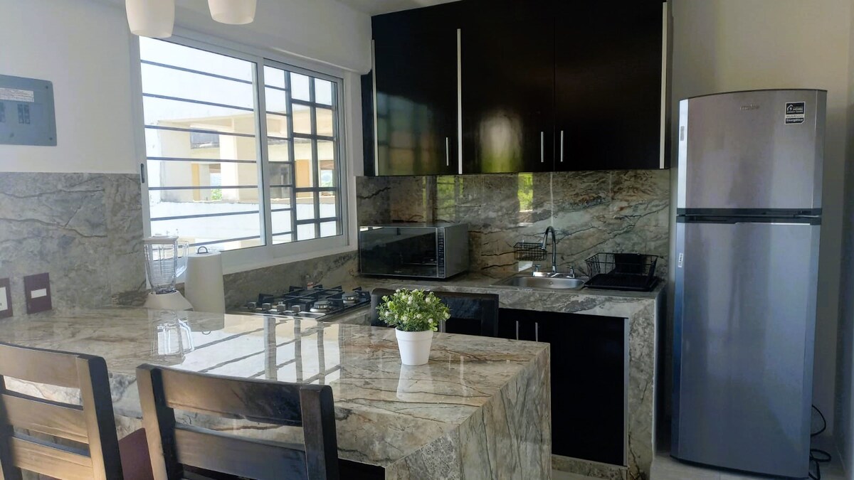 A kitchen is equipped with modern appliances, including a refrigerator and microwave. A stove with multiple burners is presented, along with ample counter space and seating at a bar with high chairs. Natural light illuminates the space through a large window.