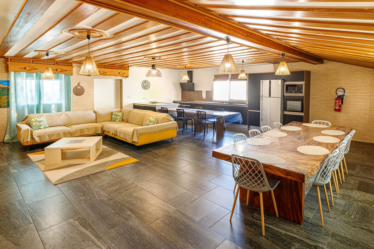 An open living area is highlighted by a spacious sectional sofa and a large dining table with seating for ten. The kitchen, equipped with modern appliances, is visible in the background. Warm wood beams and natural light create an inviting ambiance.