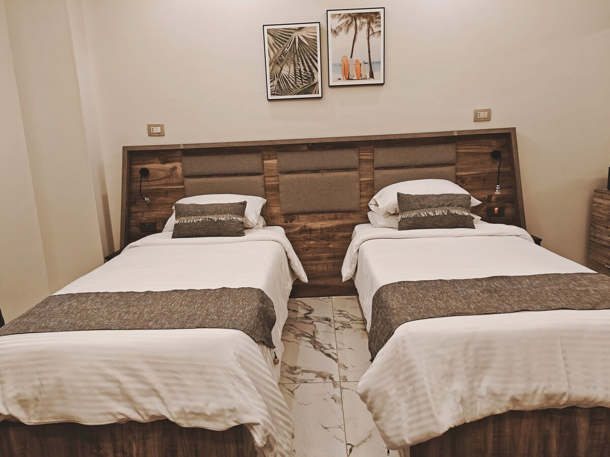 Two spacious beds are arranged side by side, each dressed in white linens and accented with light gray throws. Framed tropical prints adorn the wall above the beds, adding a touch of decor to the neutral-toned room.