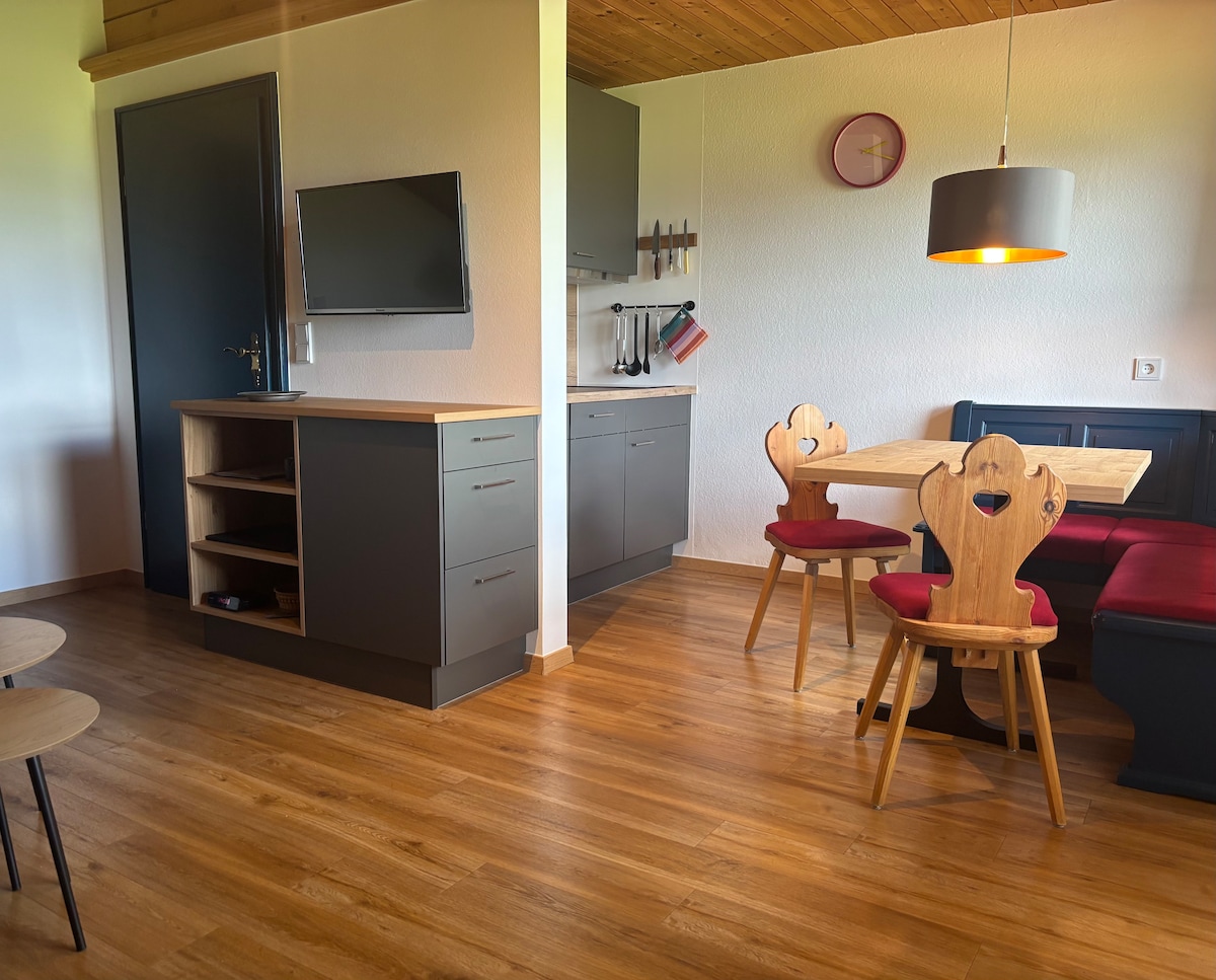 A cozy dining area features a wooden table with heart-shaped cutouts in the chairs, complemented by red upholstery. The kitchenette is equipped with modern appliances, and a flat-screen TV is mounted on the wall. Natural light fills the space, enhancing the warm wooden accents.