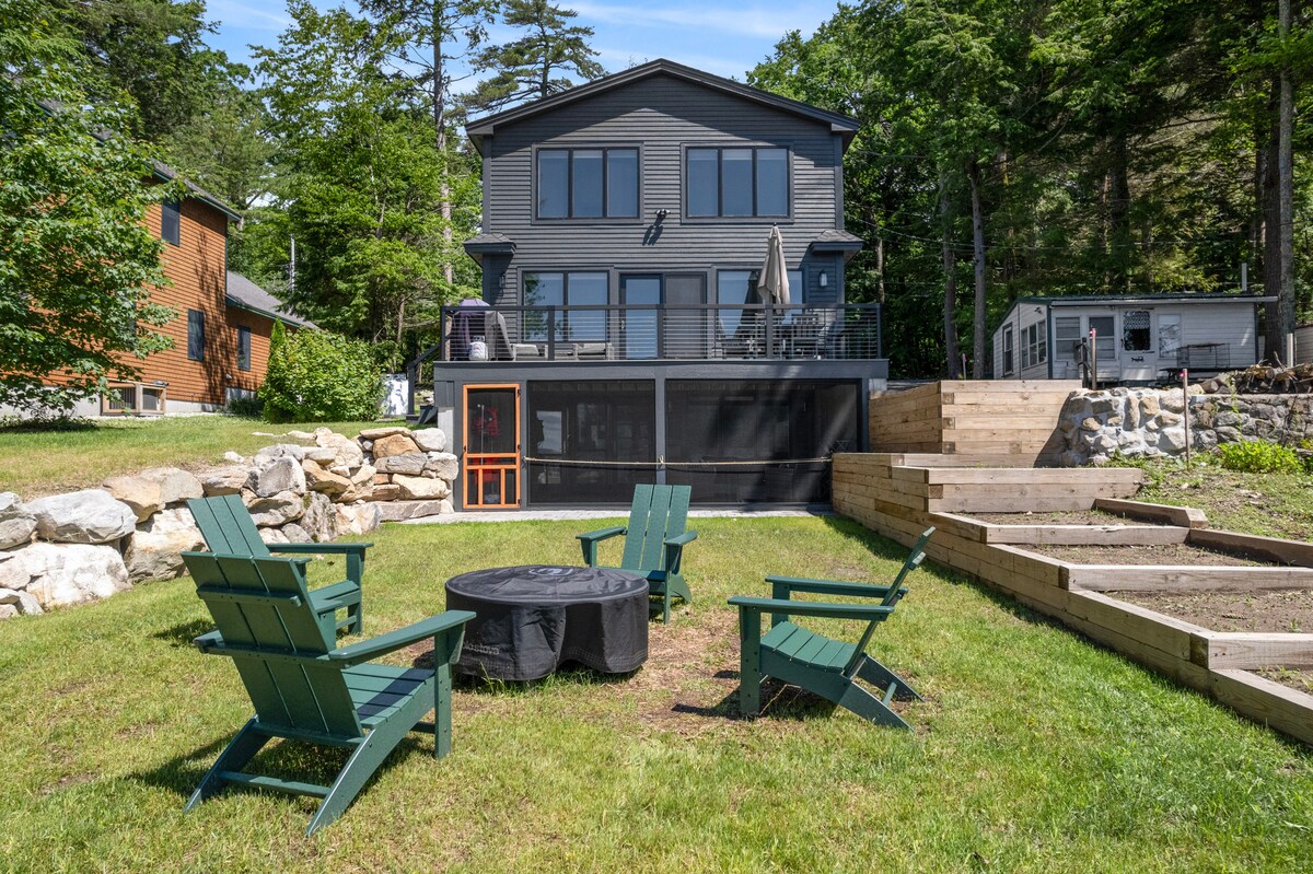 A spacious outdoor area is featured with several green adirondack chairs arranged around a black fire pit. The modern two-story home, with large windows and a screened-in patio, is visible in the background, surrounded by greenery and stone landscaping.