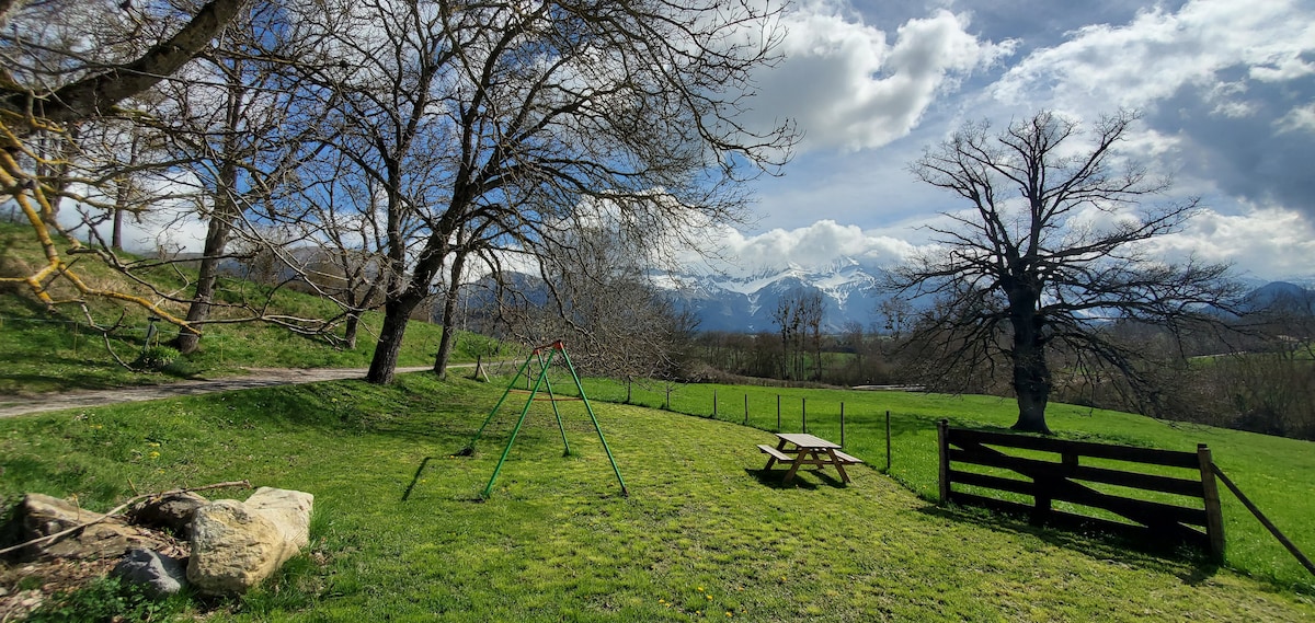 A spacious outdoor area is surrounded by trees and lush greenery, featuring a swing set and a picnic table. In the background, mountains rise against a partly cloudy sky, providing a scenic view that enhances the natural setting.