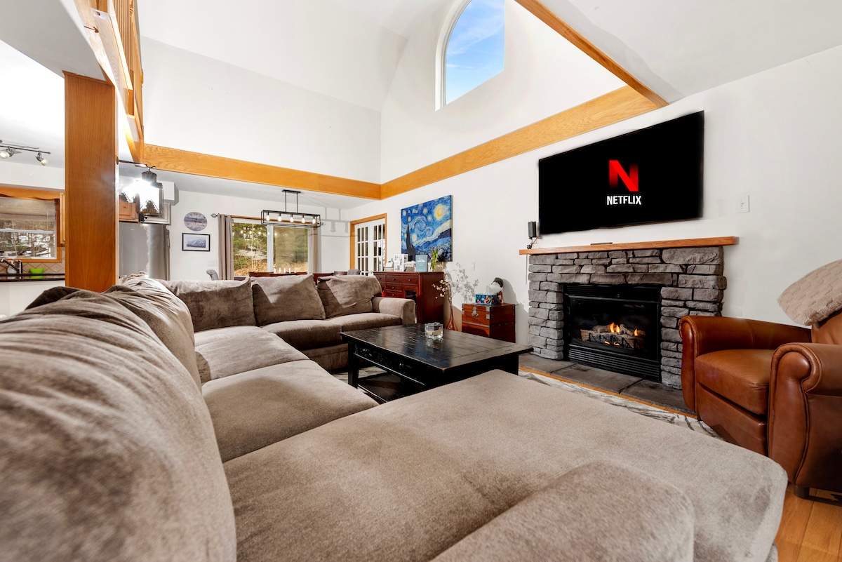 A spacious living area features a large sectional sofa arranged around a coffee table, with a stone fireplace providing warmth. A flat-screen TV is mounted above the fireplace. Large windows allow natural light to fill the room, enhancing the inviting atmosphere.