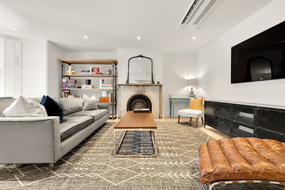 A modern living room features a comfortable sofa paired with a textured area rug. A sleek coffee table sits centered, while a bookshelf holds a variety of books. A decorative mirror is displayed above a decorative marble fireplace, and a mounted television is present on the wall.