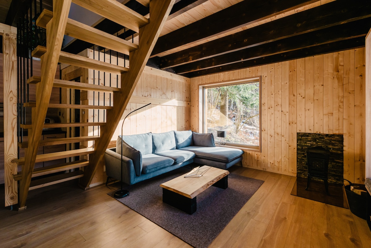 A wooden interior features a light blue sectional sofa positioned around a low coffee table on a textured rug. A window offers views of the surrounding forest, while a black wood-burning stove adds warmth to the space. Stairs lead to the upper level.