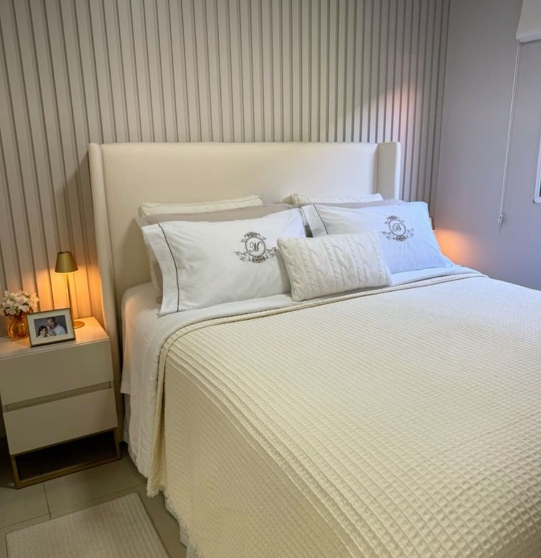 A spacious bed is dressed in a soft white coverlet, complemented by two decorative pillows. Soft lighting is provided by a bedside lamp, and a small framed photo and vase sit atop a minimal nightstand next to the bed. Neutral tones create a calm ambiance.
