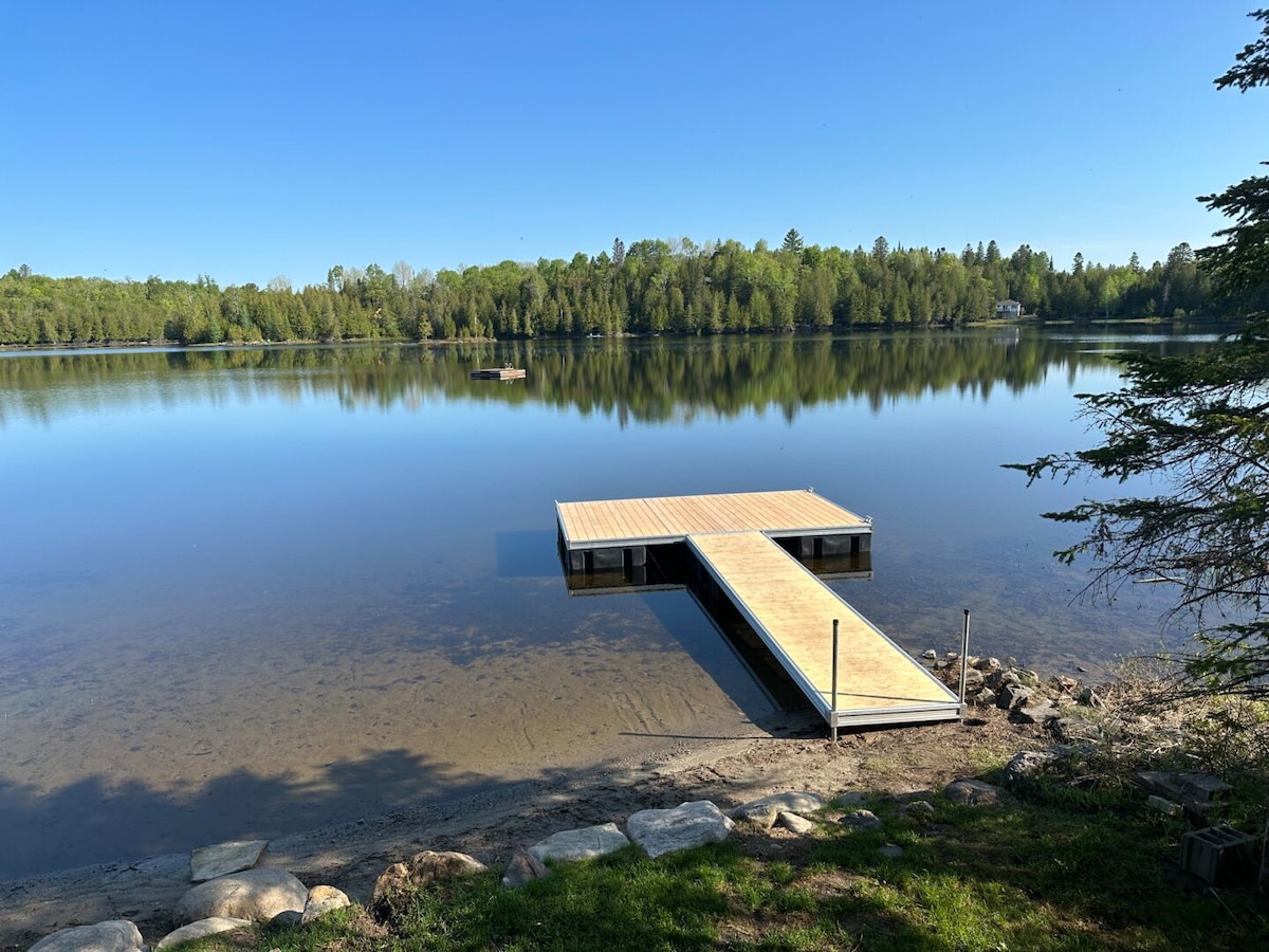 A large floating dock extends into the calm waters of a lake, surrounded by lush greenery and a clear blue sky. The serene landscape offers a peaceful atmosphere, with reflections of trees visible on the water's surface.