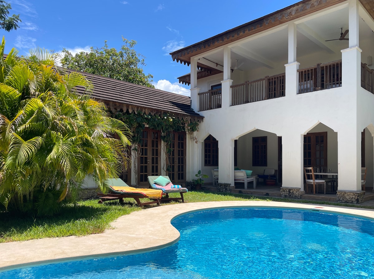 An outdoor view of the villa features a clear swimming pool surrounded by a well-maintained garden. Lounge chairs are positioned near the pool, and the two-story building displays arched openings and a balcony. Sunlight enhances the inviting atmosphere of this outdoor space.