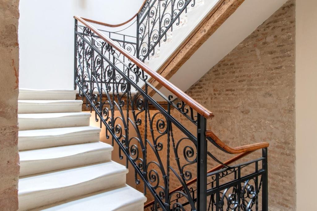 A modern staircase is highlighted, featuring elegant wrought iron railings with intricate designs. The steps are made of light-colored stone, leading upwards. The walls are adorned with a subtle texture, and natural light filters in, creating a warm ambiance in the space.