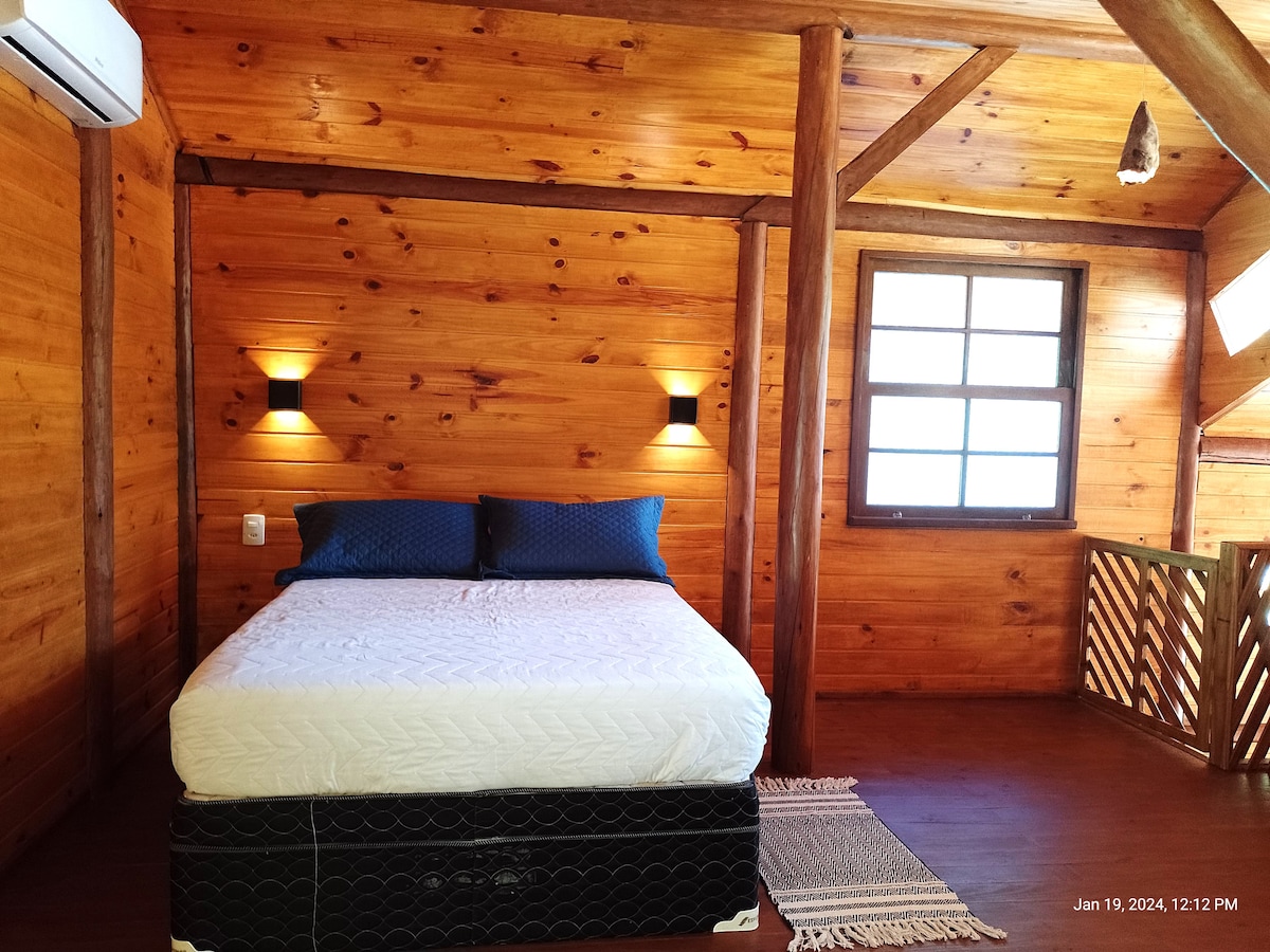 A wooden-paneled room features a spacious bed adorned with blue accent pillows. Soft lighting from wall sconces complements the natural wood tones. A window allows natural light to fill the space, providing a cozy and inviting environment.