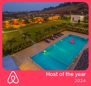 An aerial view of the villa showcases a spacious private pool surrounded by lounge chairs. Lush greenery and pathways lead to the rustic cottages in the background, softly illuminated by warm lights, highlighting the serene outdoor atmosphere of the property.