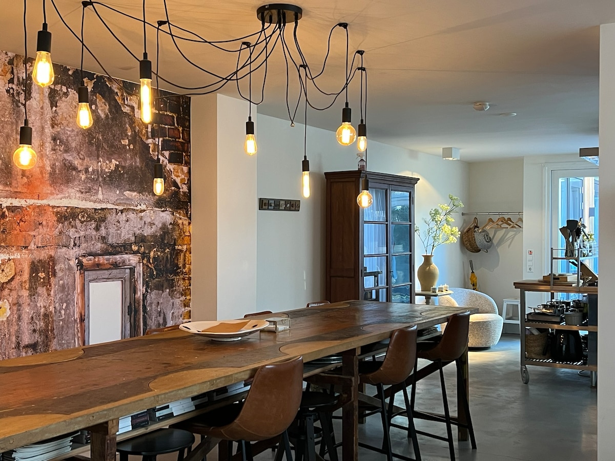 A spacious dining area is showcased, featuring a long, rustic wooden table surrounded by several high-backed chairs. Industrial-style pendant lights hang overhead, illuminating the room. A brick wall adds texture, while a cabinet and decorative elements contribute to the inviting design.