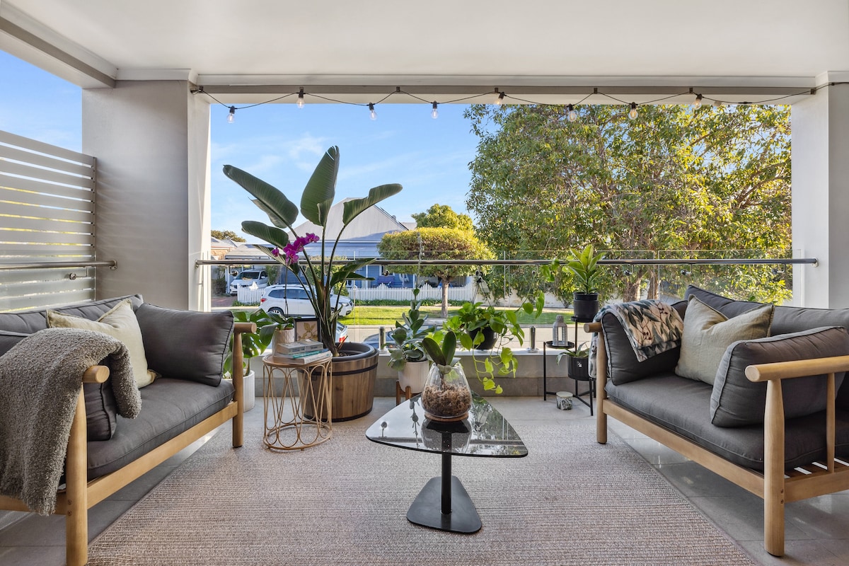 A stylish balcony area features two comfortable lounge chairs and a central glass table. Lush greenery and potted plants add a touch of nature, while warm ambient lighting and street views create a serene outdoor environment.