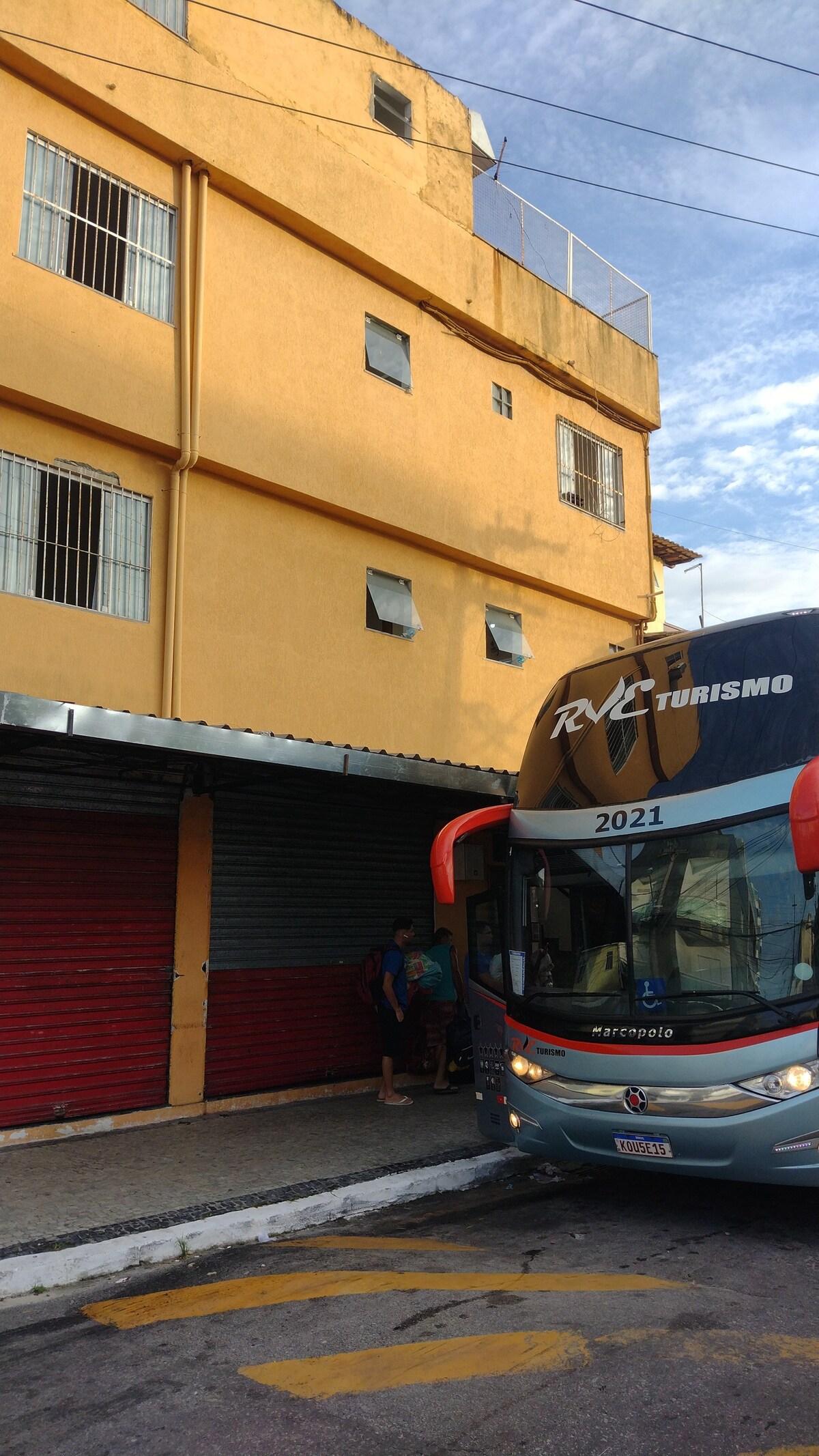 A brightly painted yellow building is depicted with large windows and a metal awning. A bus is parked in front, featuring a logo that reads 'RNC Turismo.' Nearby, individuals are seen interacting outside the building, with a view of a partially cloudy sky above.