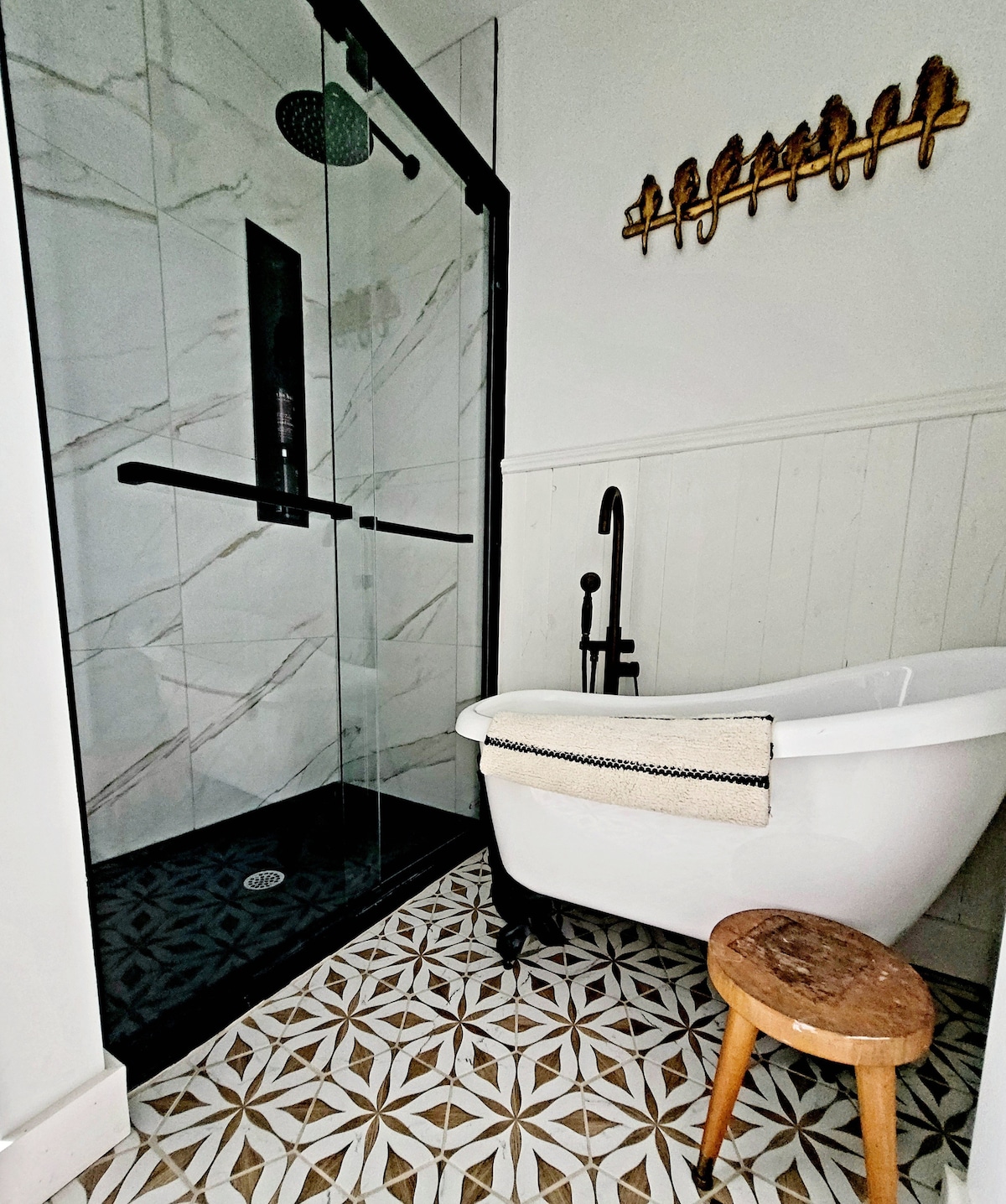 A modern bathroom features a clear glass shower with a sleek black frame and a rainfall showerhead. A small, white bathtub is positioned beside a wooden stool, while intricate tile patterns adorn the floor, adding visual interest to the space.