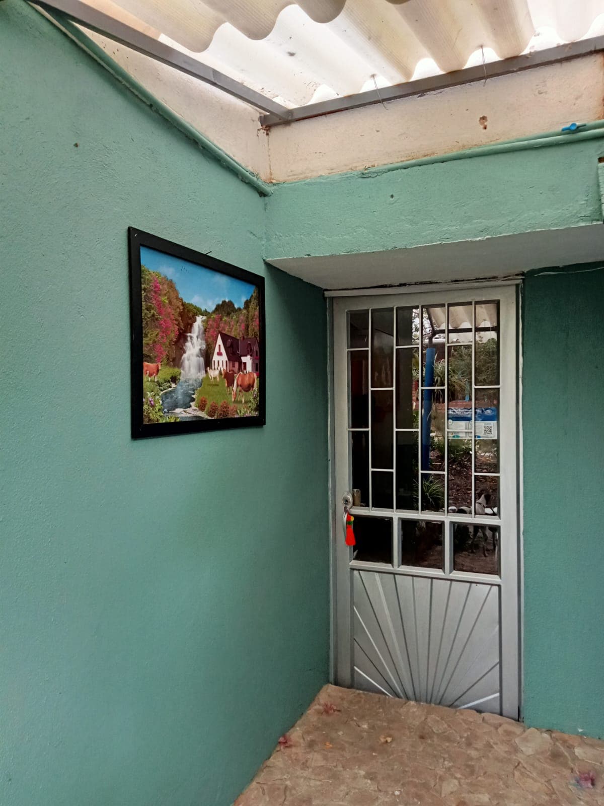 An entrance is highlighted by a light gray door, featuring a decorative metal grille. A colorful landscape painting adorns the green wall, contributing a vibrant touch. Natural light is noted, illuminating the inviting entryway.