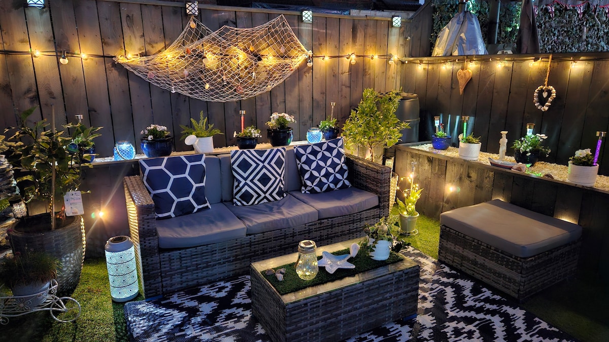 A cozy outdoor seating area features a comfortable sofa set with decorative cushions and a cushioned ottoman. Soft lighting from fairy lights and lanterns enhances the ambiance, complemented by potted plants and vibrant decorations on the surrounding walls.