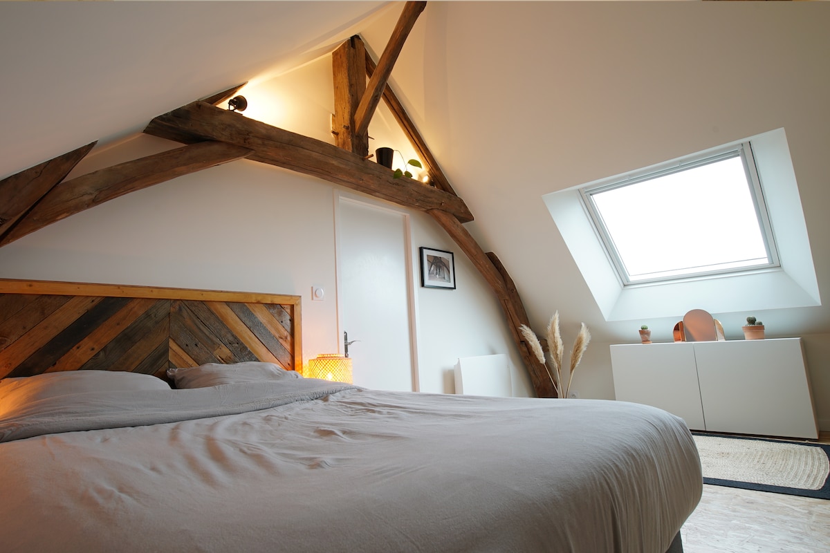 The bedroom features a double bed with a neutral-toned duvet and artistic wooden headboard. Exposed wooden beams accentuate the sloped ceiling, while a skylight provides natural light. A minimalist dresser and decorative plants are visible, enhancing the cozy atmosphere.