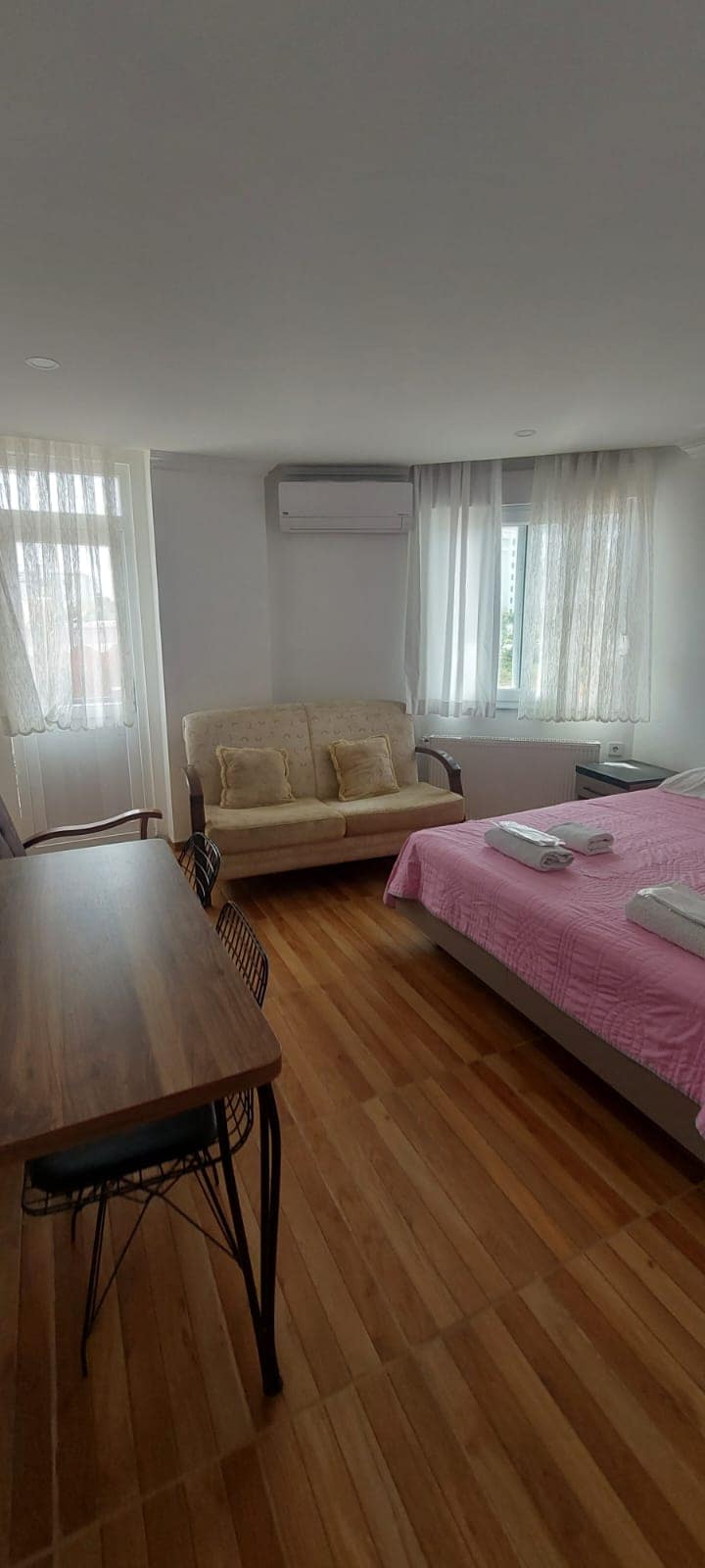 A bright and airy room features two large windows with sheer curtains, allowing ample natural light. A spacious bed adorned with a pink coverlet is accompanied by neatly arranged towels. A cozy seating area with a light-colored couch sits beside a wooden table and chair.