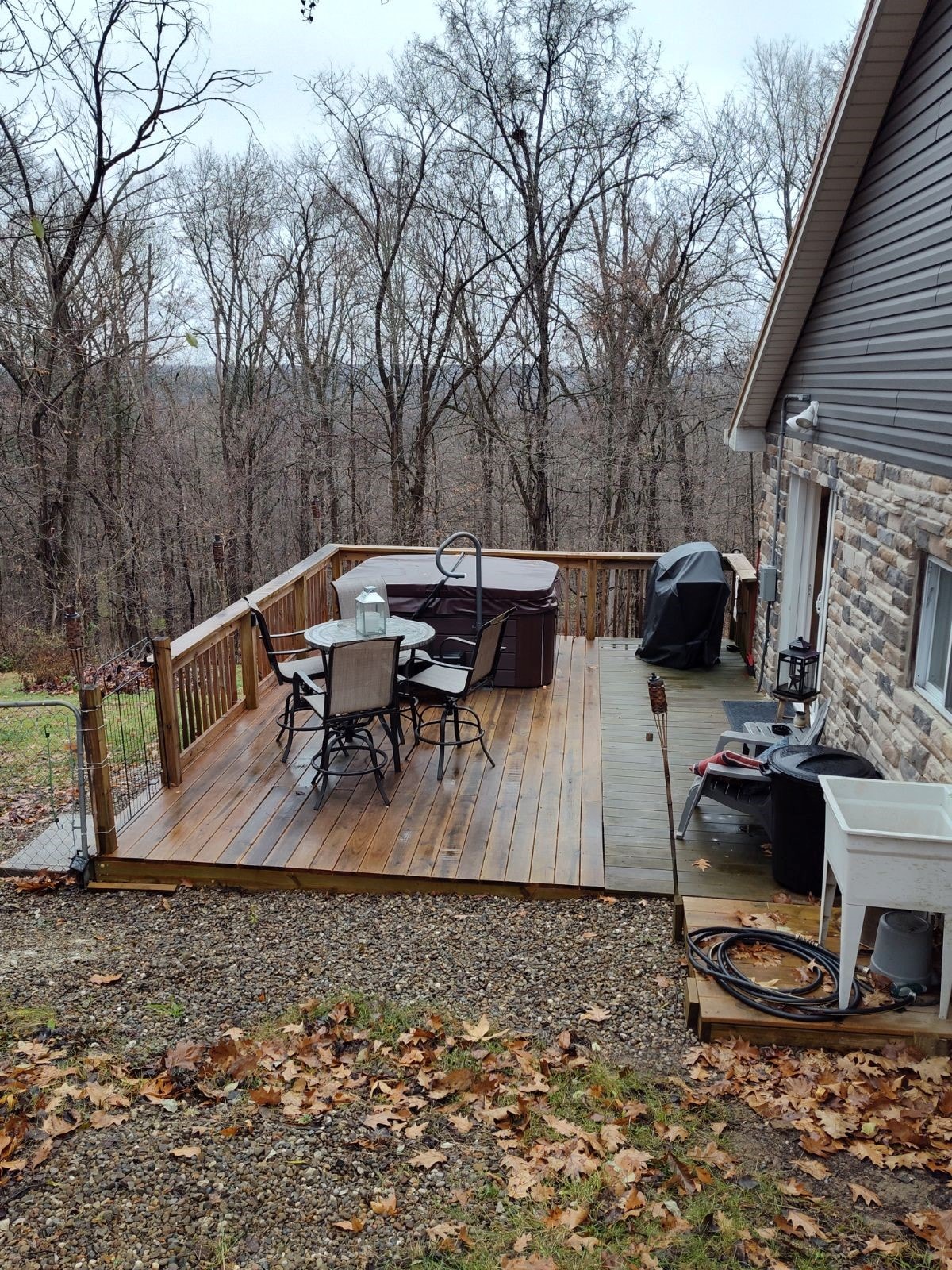 A spacious deck features a hot tub, dining table with chairs, and a charcoal grill, providing a perfect outdoor setting. The deck is surrounded by trees in a serene, wooded area, showcasing a natural atmosphere. Fallen leaves cover the ground, indicating the season.