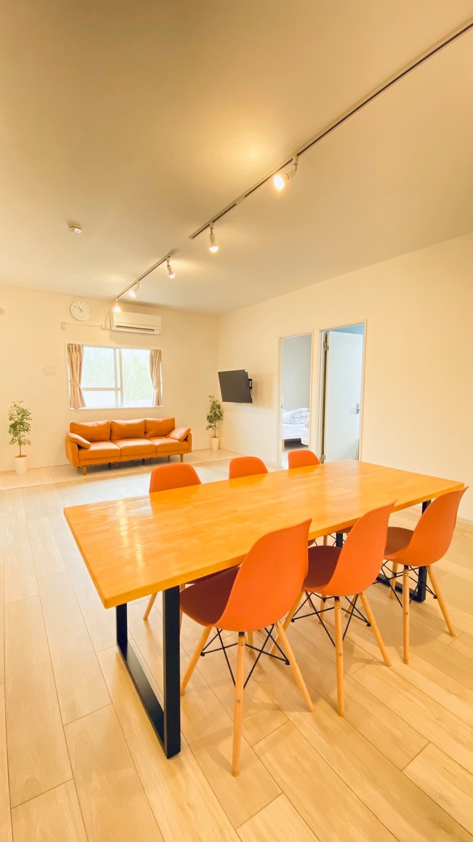 An open interior space features a dining table with six chairs, complemented by a cozy sofa in the background. Soft lighting enhances the neutral-colored walls, while a window allows natural light to brighten the area. A television is mounted on the wall, providing entertainment options.