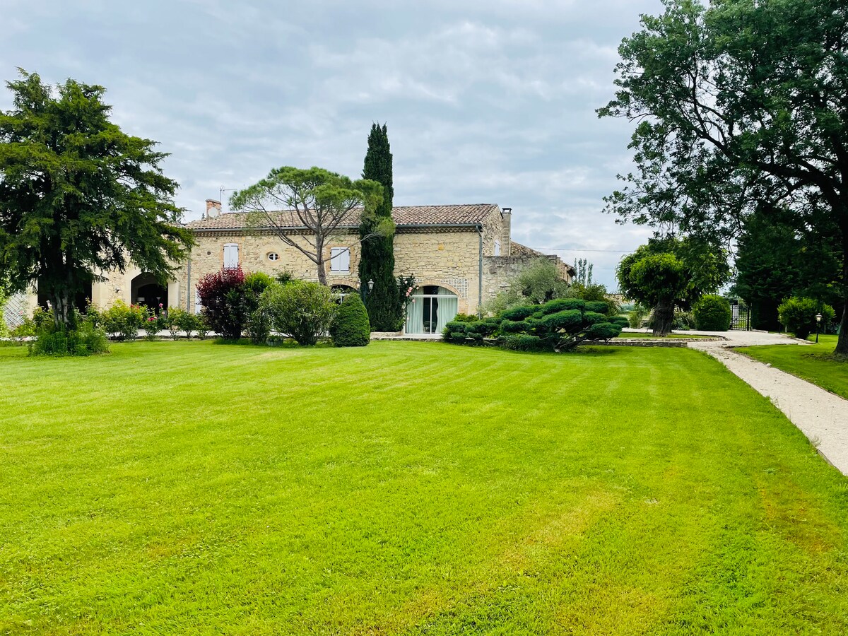 The exterior of a historic Provençal mas is visible, surrounded by manicured lawns and lush greenery. A winding pathway leads to the entrance, with various trees and shrubs enhancing the landscape. The serene atmosphere is complemented by a partly cloudy sky.