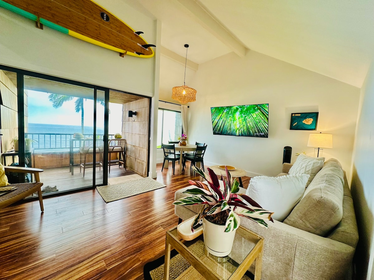 A bright living area features a spacious white sofa and a decorative coffee table with plants. A dining space with a table for four sits nearby, while large glass doors open to a private lanai. Natural light fills the room, highlighting the wooden flooring.