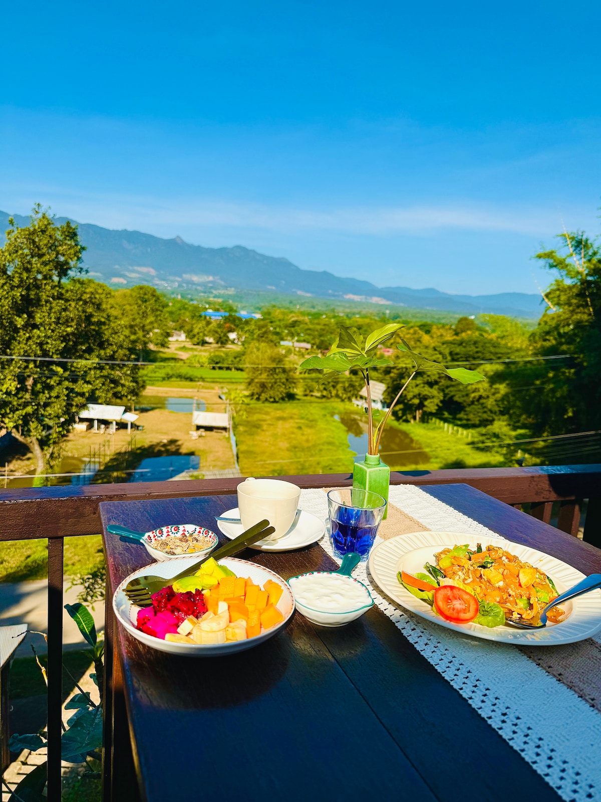 A table is set on a balcony, displaying a colorful array of sliced fruits and a meal alongside a drink and a cup. The backdrop features expansive views of green fields and distant mountains under a clear blue sky.