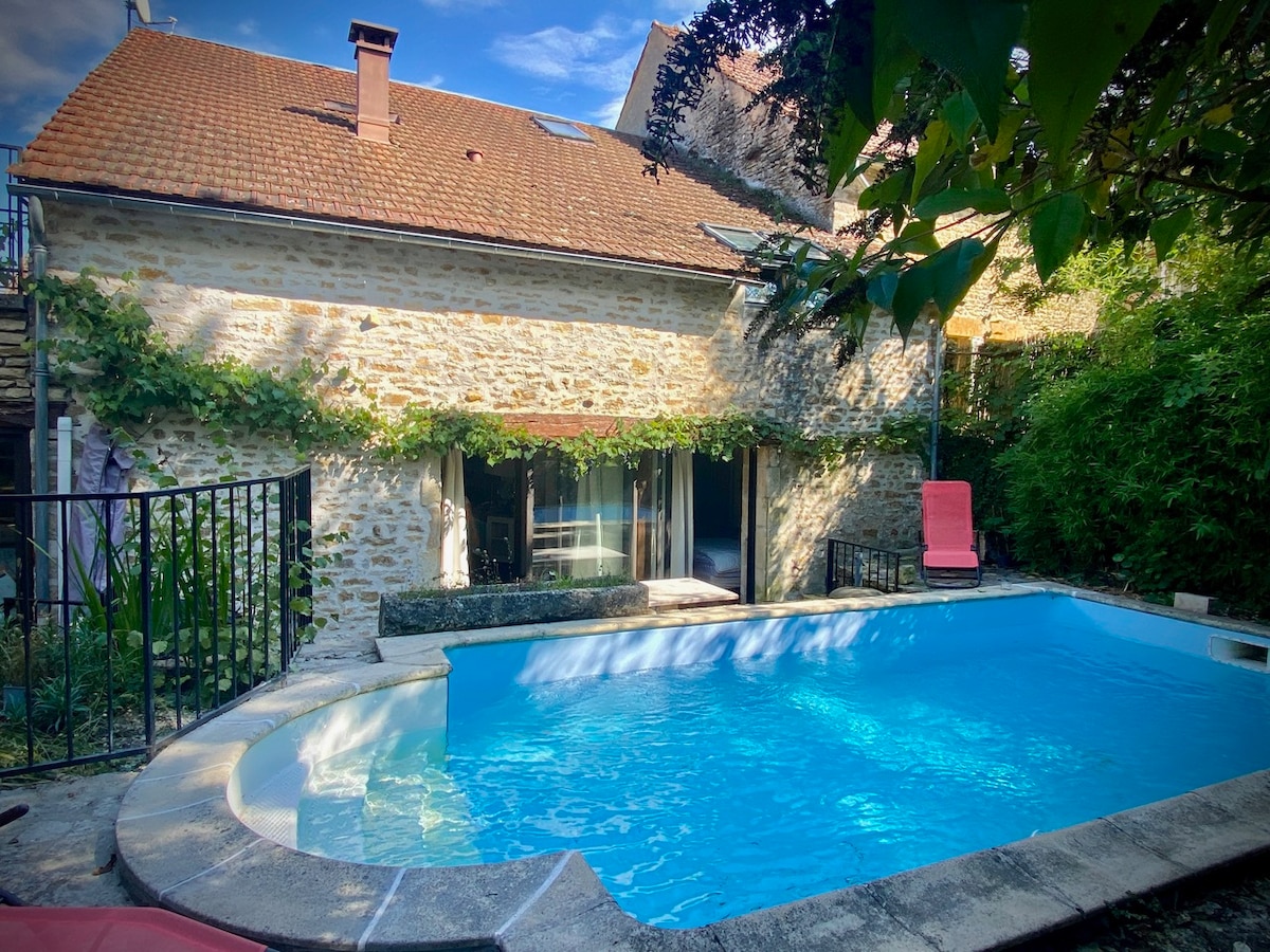 A secluded outdoor area features a private pool with clear blue water, surrounded by a stone wall. A terrace is visible in front of the house, with large windows offering light inside. Lush greenery and climbing plants add to the serene atmosphere.