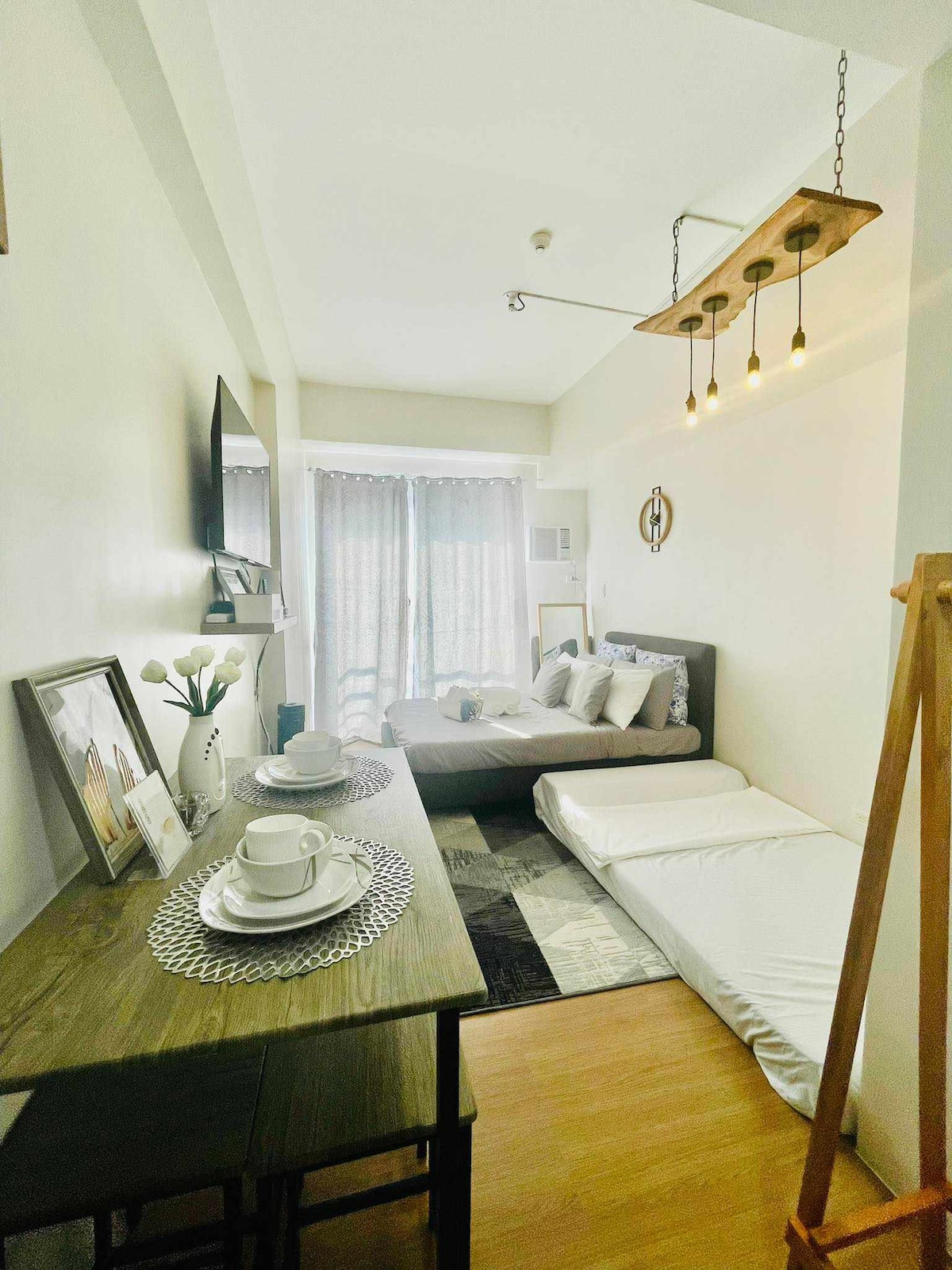 A well-arranged studio space is visible, featuring a spacious bed with decorative pillows and a rollaway mattress. A dining table is set with dishware, while a smart TV and window draped with light curtains complement the area, enhancing the overall comfort of the setting.