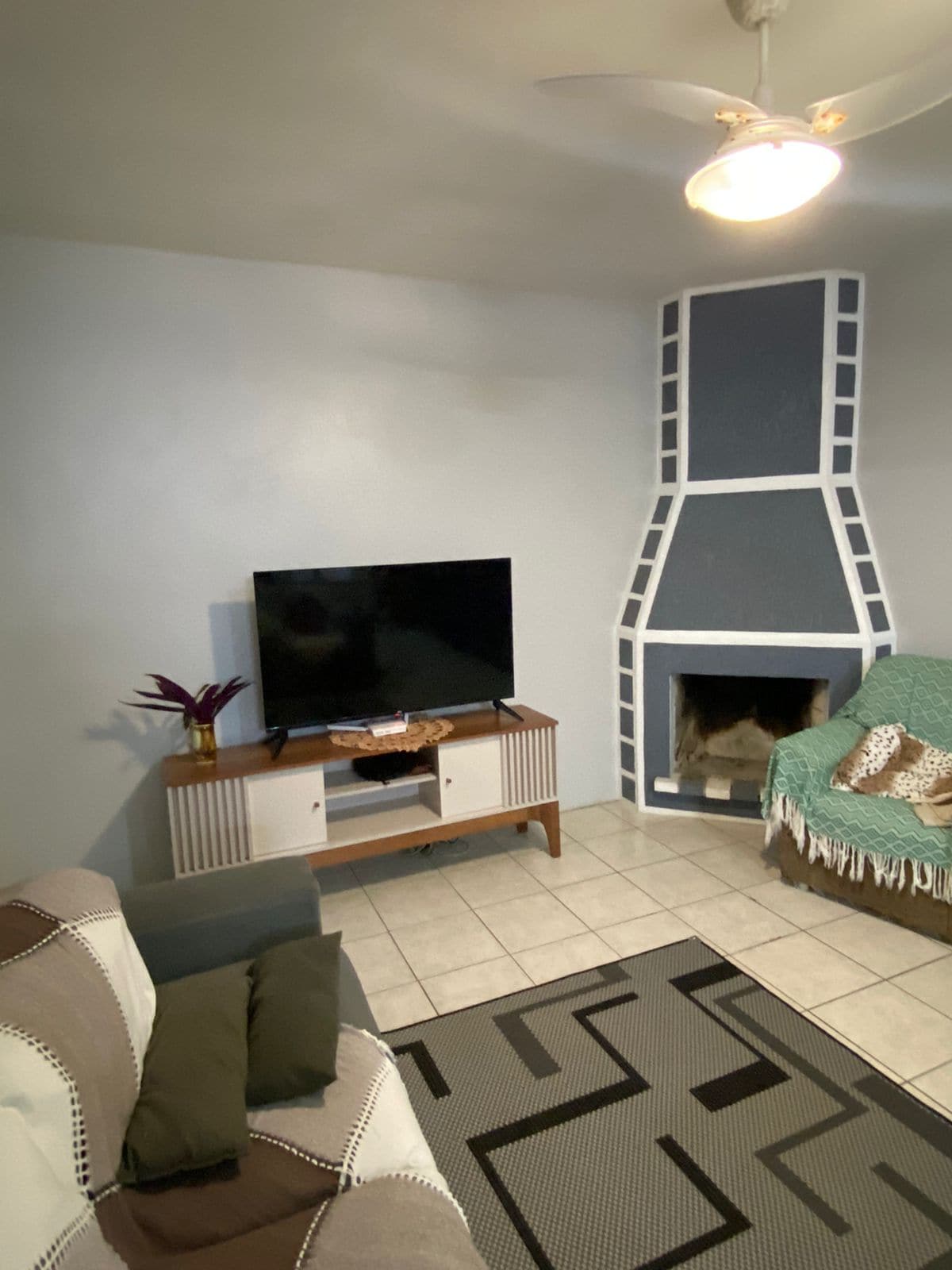 A cozy living area features a flat-screen television atop a wooden media console. A fireplace, accented with a modern design, sits beside a comfortable armchair draped with a textured throw. The space is enhanced by tile flooring and neutral walls.