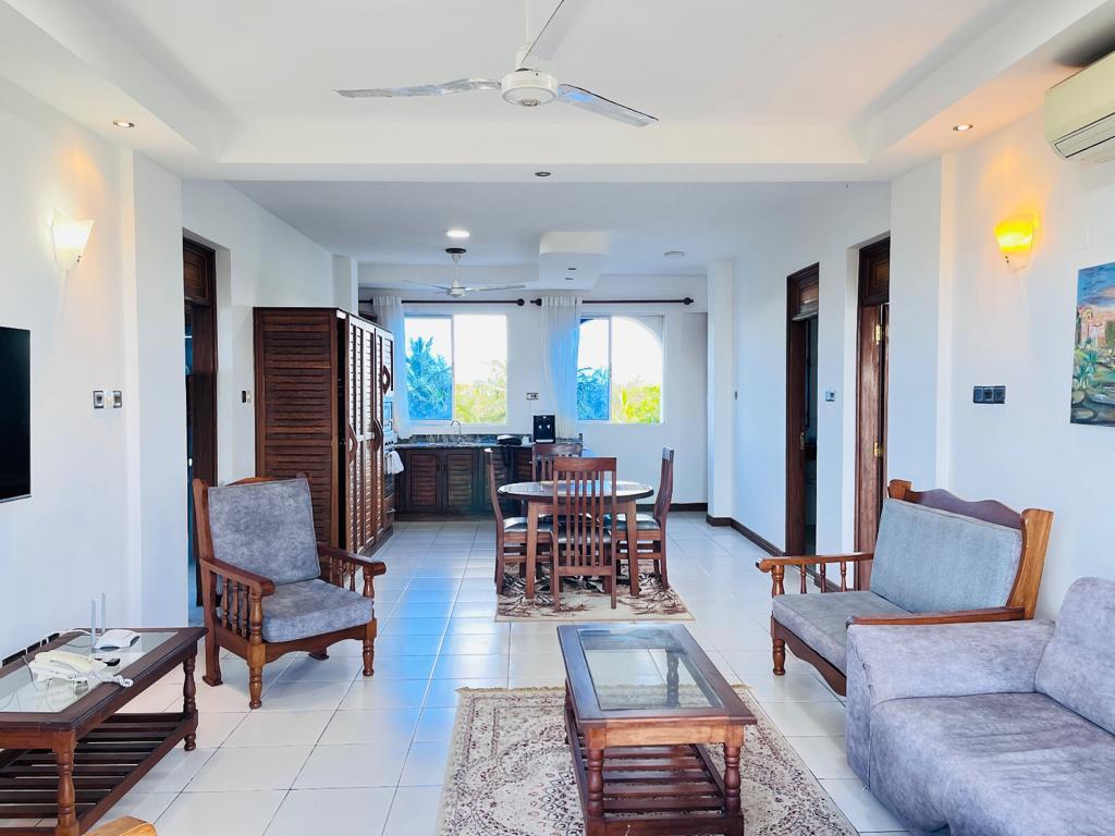 A light-filled living and dining area features wooden furniture, including a dining table with chairs and a comfortable seating arrangement. Large windows provide views of the outdoors and allow natural light to fill the space. Decorative elements include a rug and wall art.